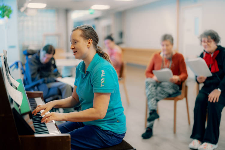 Hoitaja soittaa pianoa ja asiakkaat kuuntelevat ja laulavat mukana.